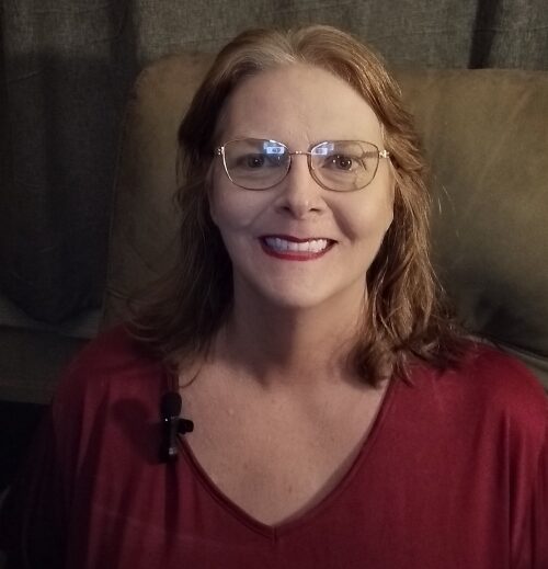 Lorelei Molinari — Speaker and Guide for Comfort in the Chaos | Comfort In The Chaos Lorelei Molinari wearing glasses and a maroon blouse, smiling with a microphone clipped to her collar against a neutral background.